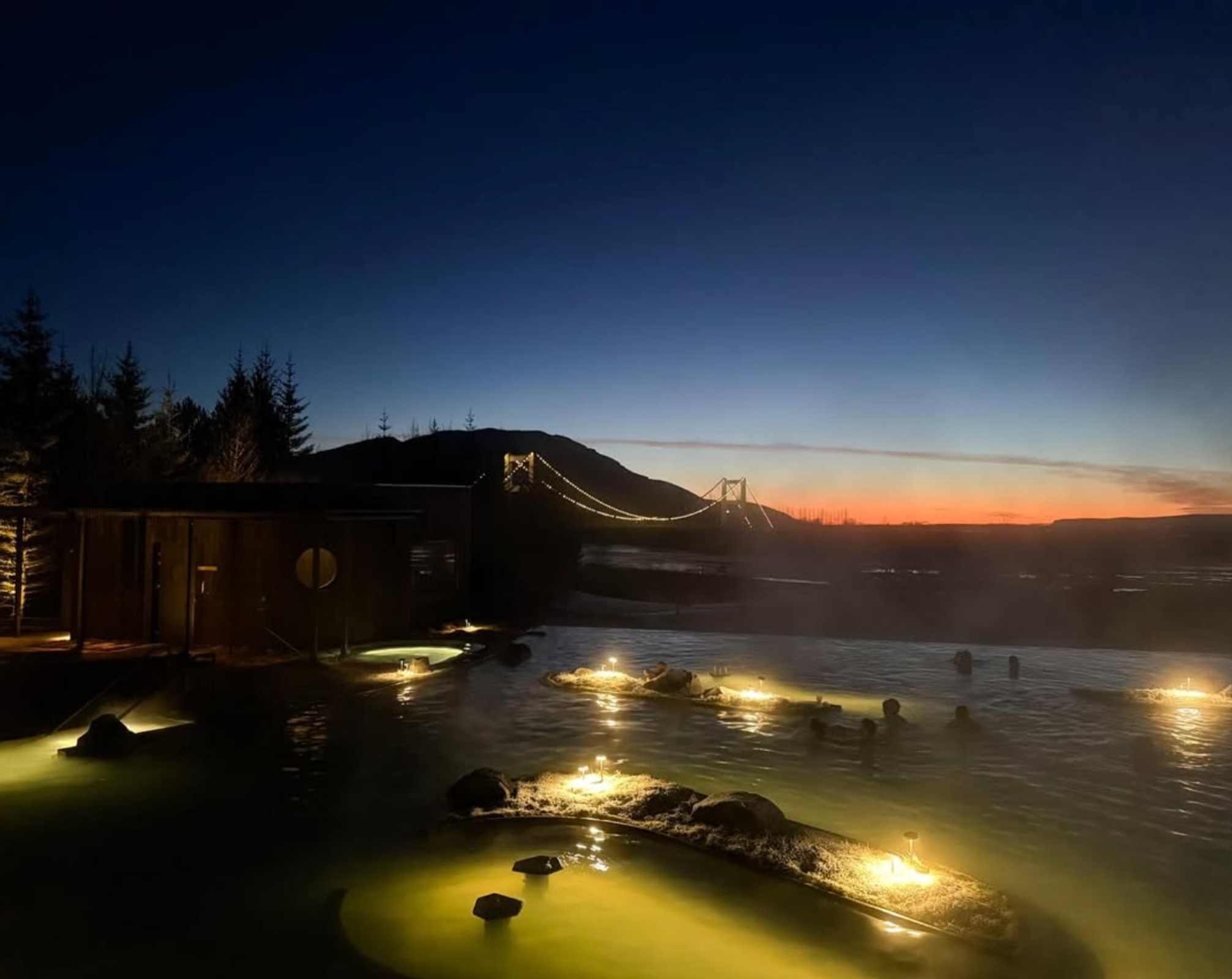 Personas bañándose en una fuente termal geotérmica luminosa al atardecer, con un puente colgante iluminado y un edificio de madera.