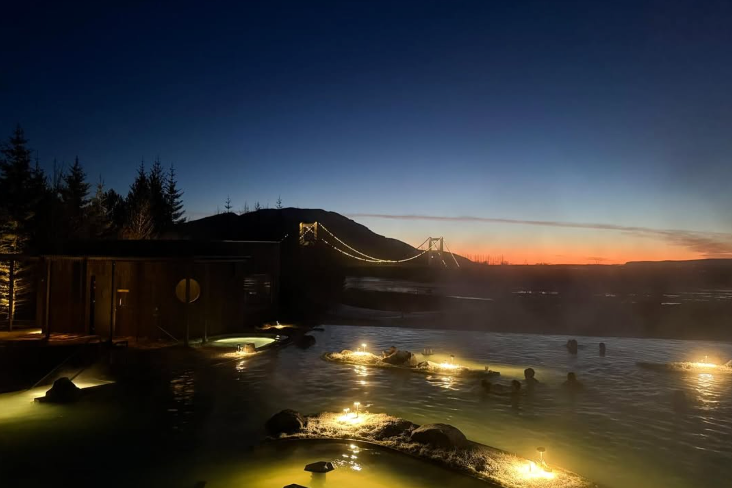 Personas bañándose en una fuente termal geotérmica luminosa al atardecer, con un puente colgante iluminado y un edificio de madera.