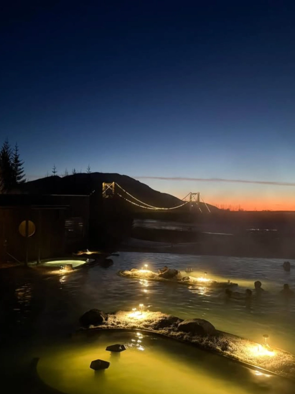 Twilight scene of people relaxing in a glowing hot spring, with a lit bridge and sunset sky in the background.