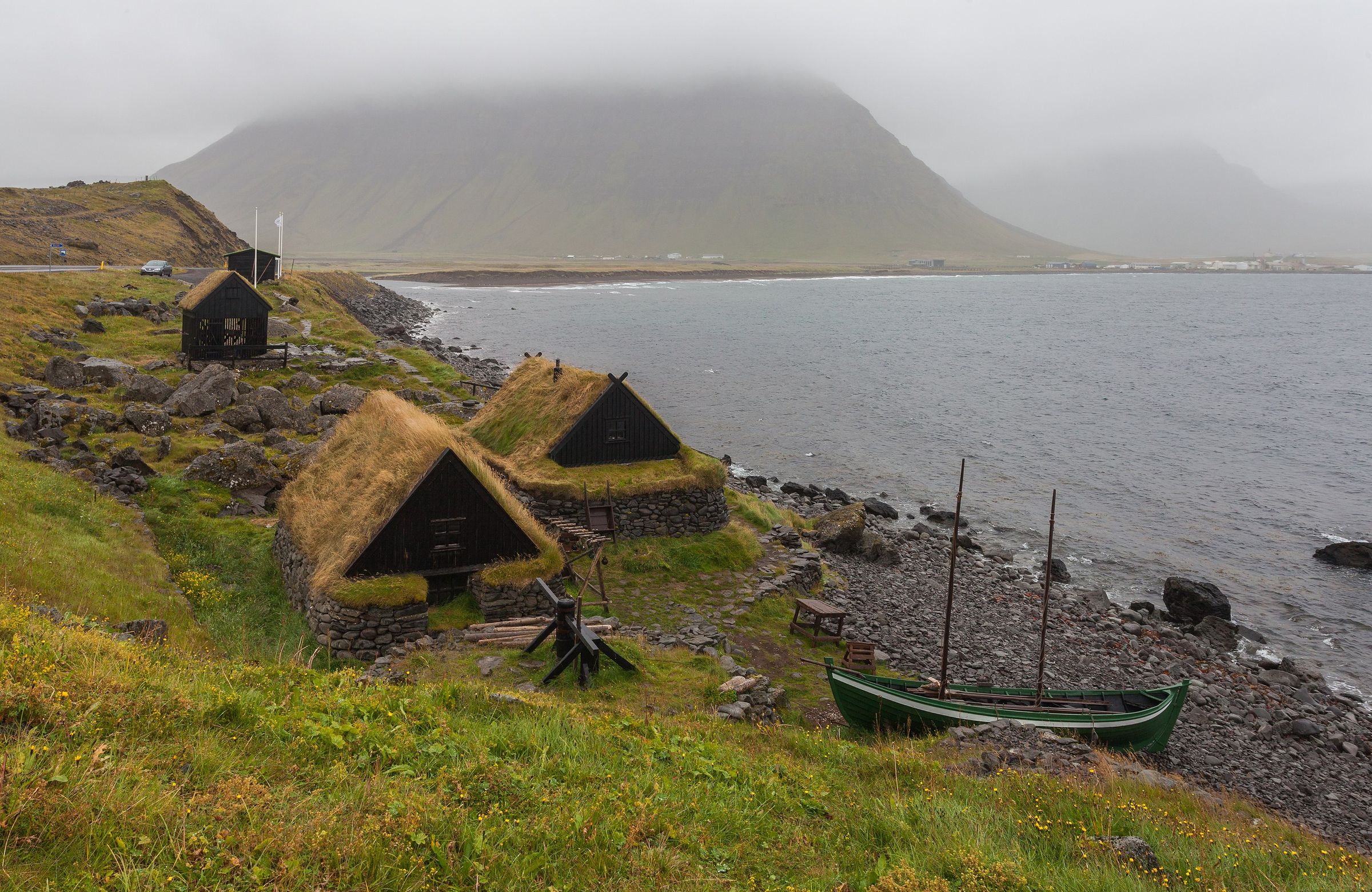 Traditional grass-roofed houses and a green boat on a rocky shore beneath a misty mountain.