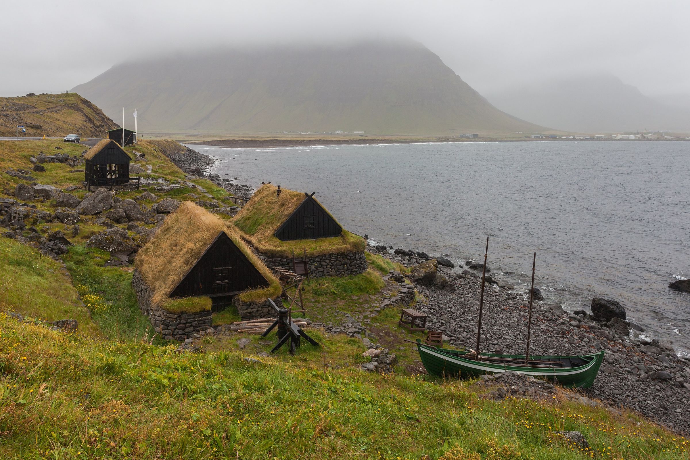 Traditional grass-roofed houses and a green boat on a rocky shore beneath a misty mountain.