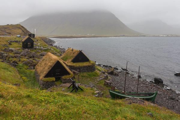 Traditional grass-roofed houses and a green boat on a rocky shore beneath a misty mountain.