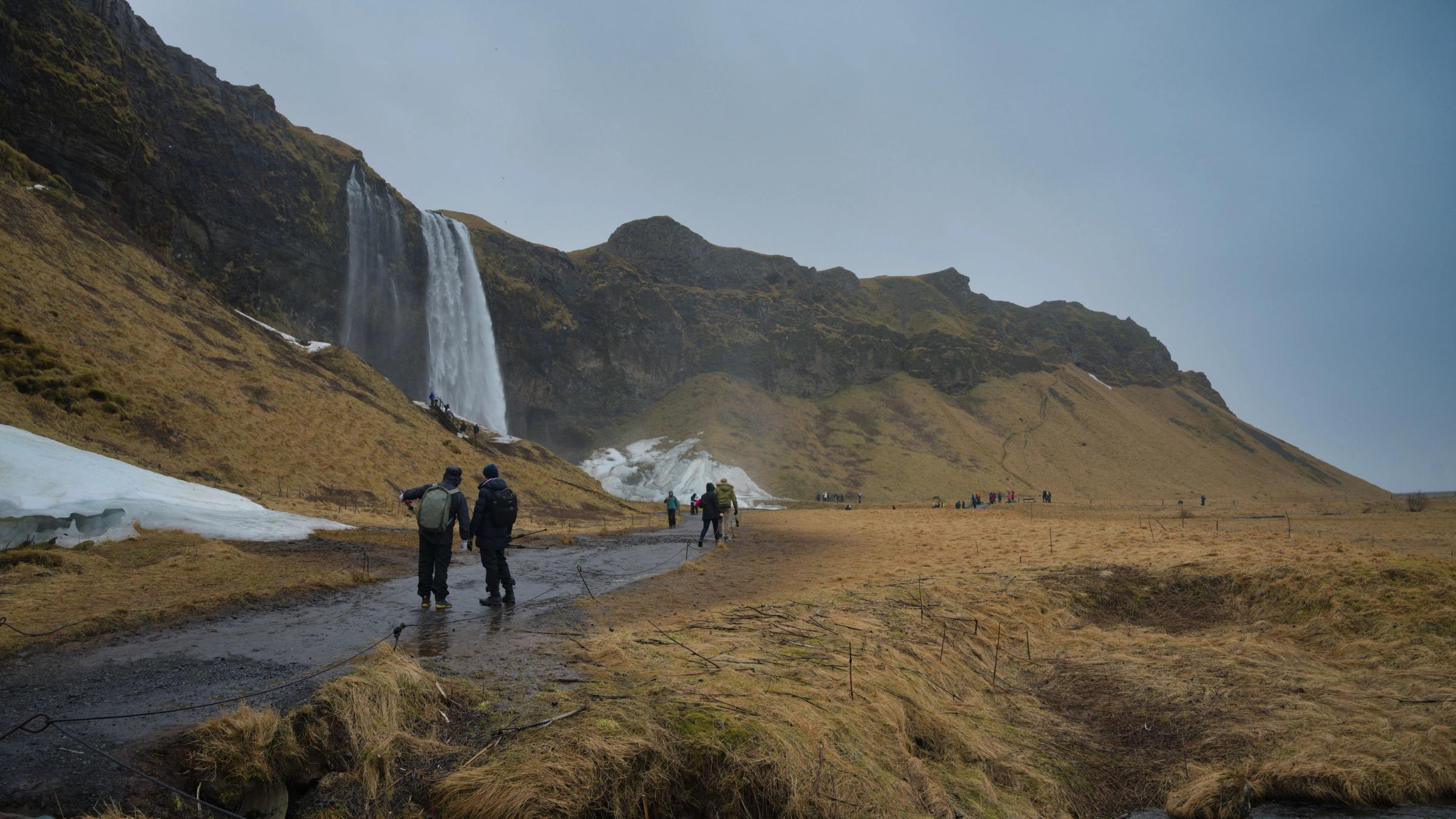 Hikers on a path approaching a large waterfall cascading from a rocky mountain, with grassy fields and patches of snow.