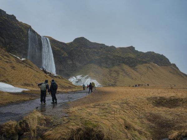 Hikers on a path approaching a large waterfall cascading from a rocky mountain, with grassy fields and patches of snow.