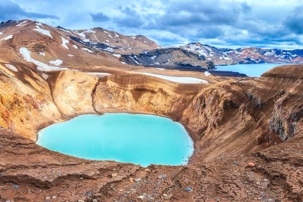Lago de cráter turquesa en un paisaje volcánico árido con montañas nevadas a lo lejos y otros lagos.