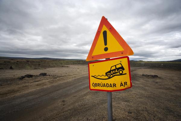 Señales de advertencia islandesas para ríos sin puentes, mostrando un 4x4 cruzando el agua, en un camino de tierra desolado.