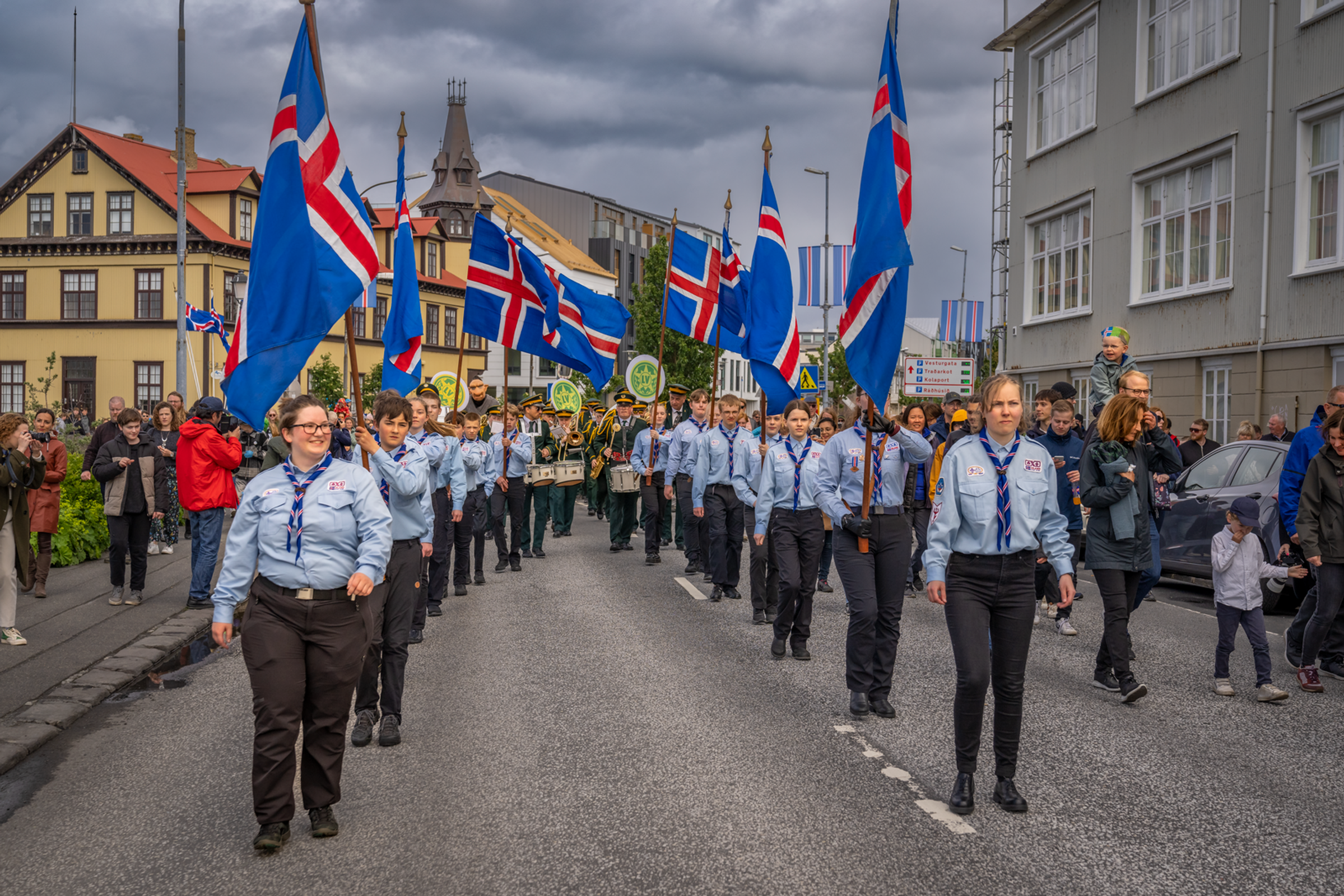 A parade of Scouts carrying Icelandic flags marches down a street lined with spectators and buildings under an overcast sky.