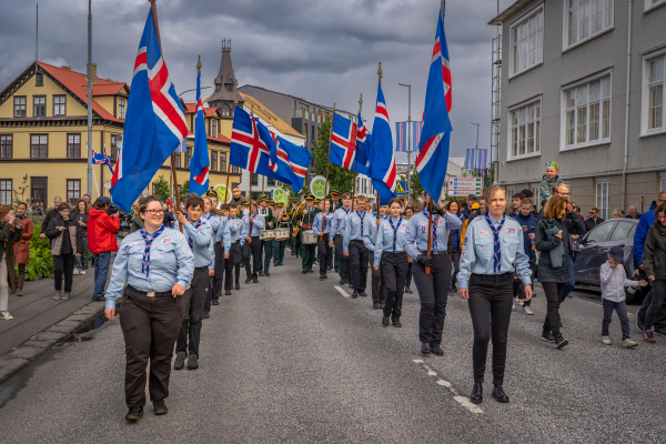 A parade of Scouts carrying Icelandic flags marches down a street lined with spectators and buildings under an overcast sky.