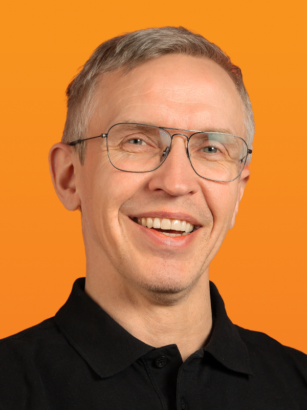 Smiling man with gray hair and glasses in a black polo shirt against an orange background.