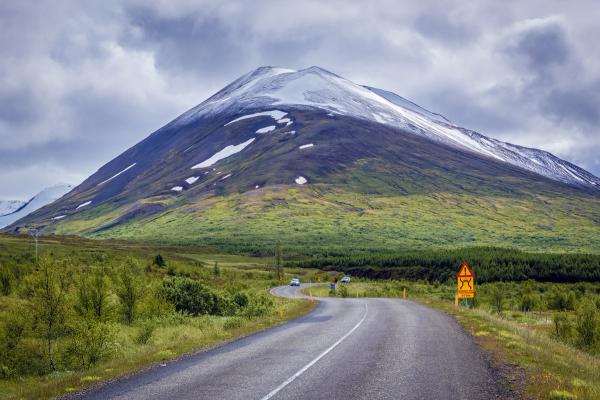 Winding road through a green landscape towards a snow-capped mountain under a cloudy sky.