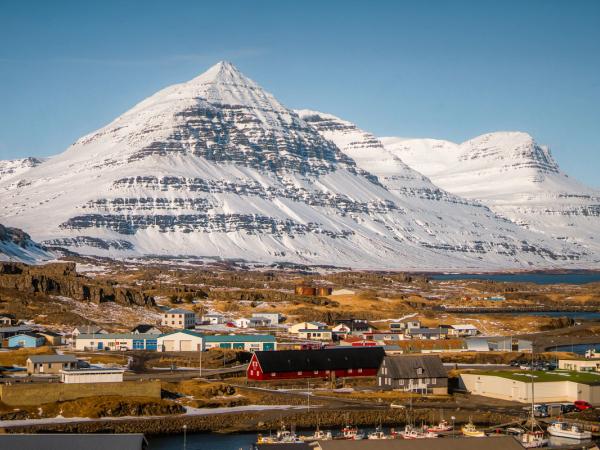 A village with a harbor sits below massive snow-capped mountains and a clear blue sky.