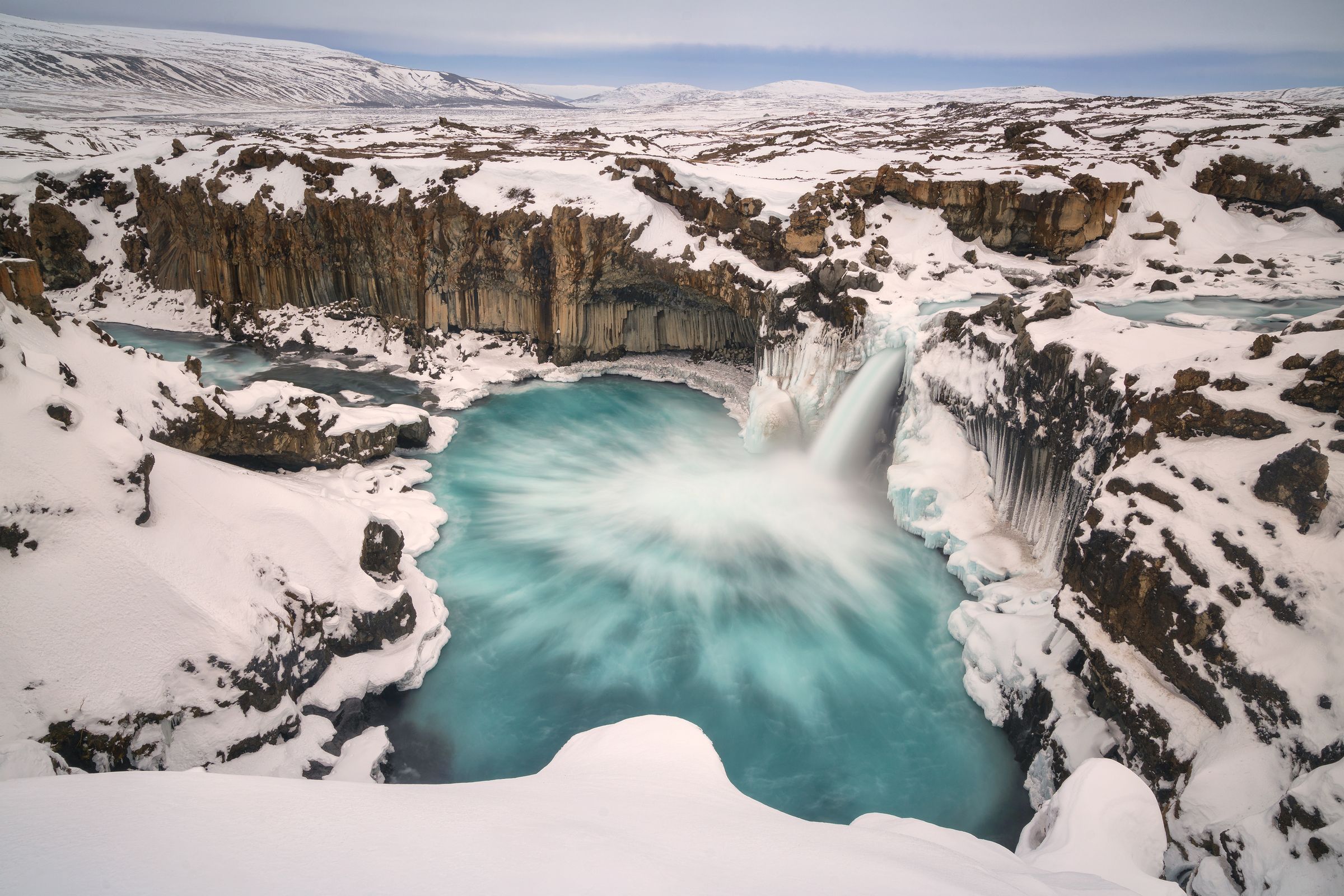 A vibrant turquoise waterfall cascades into a swirling pool in a snowy canyon with dark basalt columns.