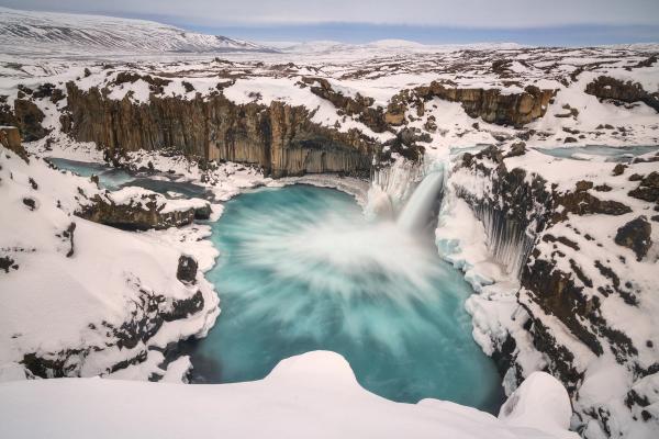 A vibrant turquoise waterfall cascades into a swirling pool in a snowy canyon with dark basalt columns.