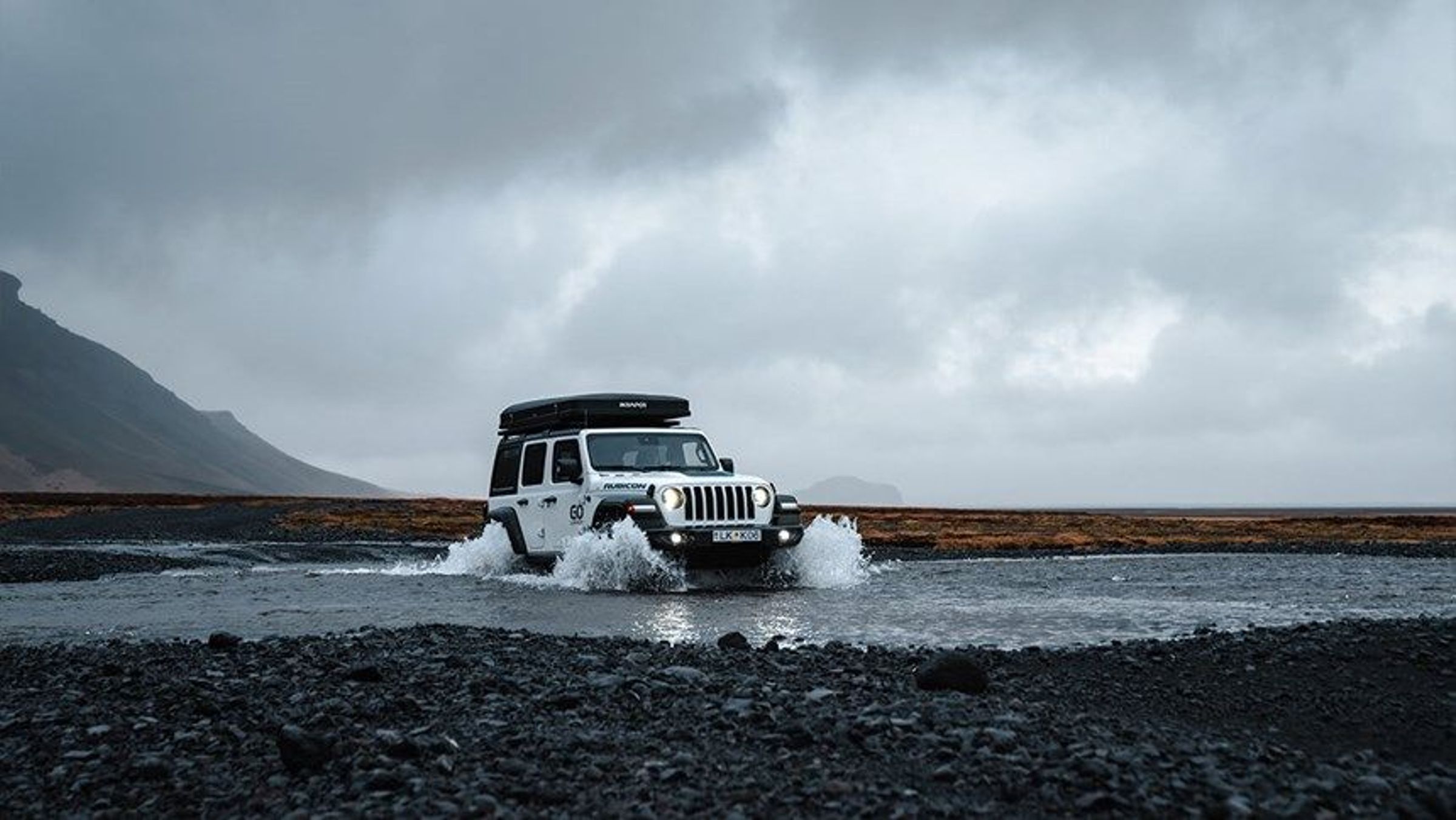 A white Jeep with a rooftop tent drives through a shallow river, splashing water in a rugged, cloudy landscape.