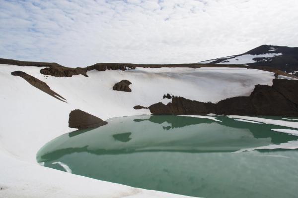 A bright turquoise lake surrounded by snow-covered brown hills under a cloudy sky.
