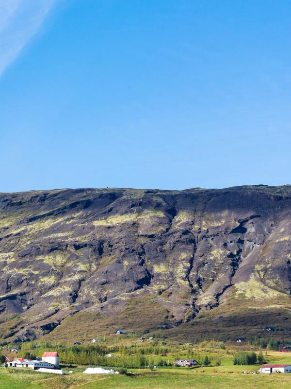 A large dark rocky mountain with green patches overlooks a green valley with scattered buildings under a clear blue sky.