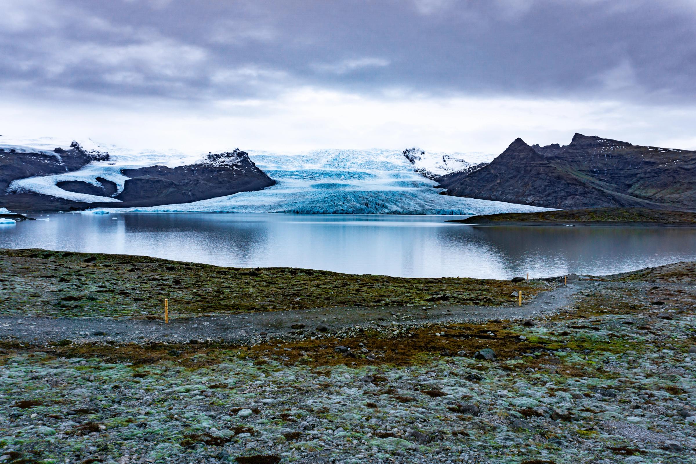 A glacial lake with a large blue and white glacier and dark mountains in the background, under an overcast sky.