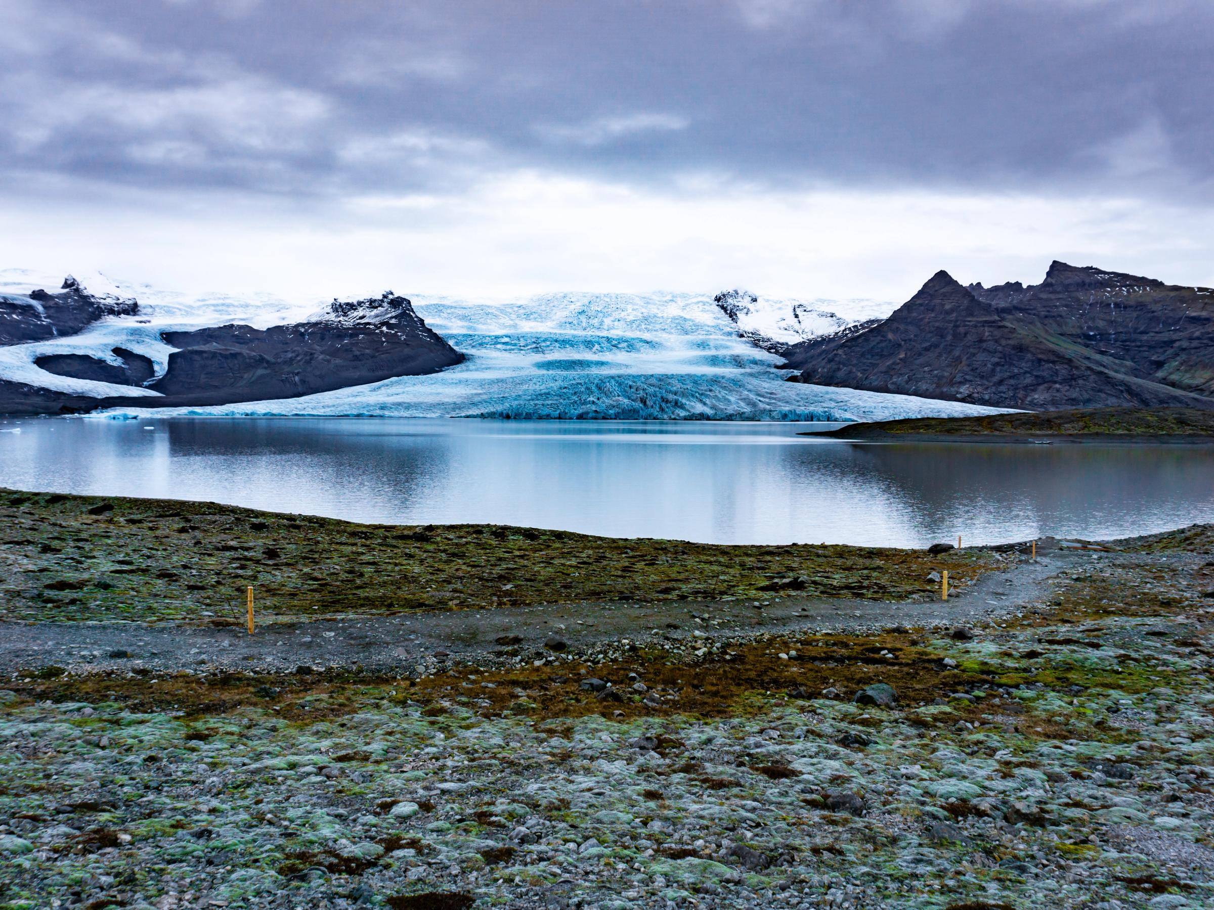 A glacial lake with a large blue and white glacier and dark mountains in the background, under an overcast sky.