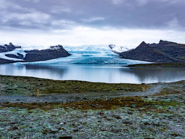A glacial lake with a large blue and white glacier and dark mountains in the background, under an overcast sky.