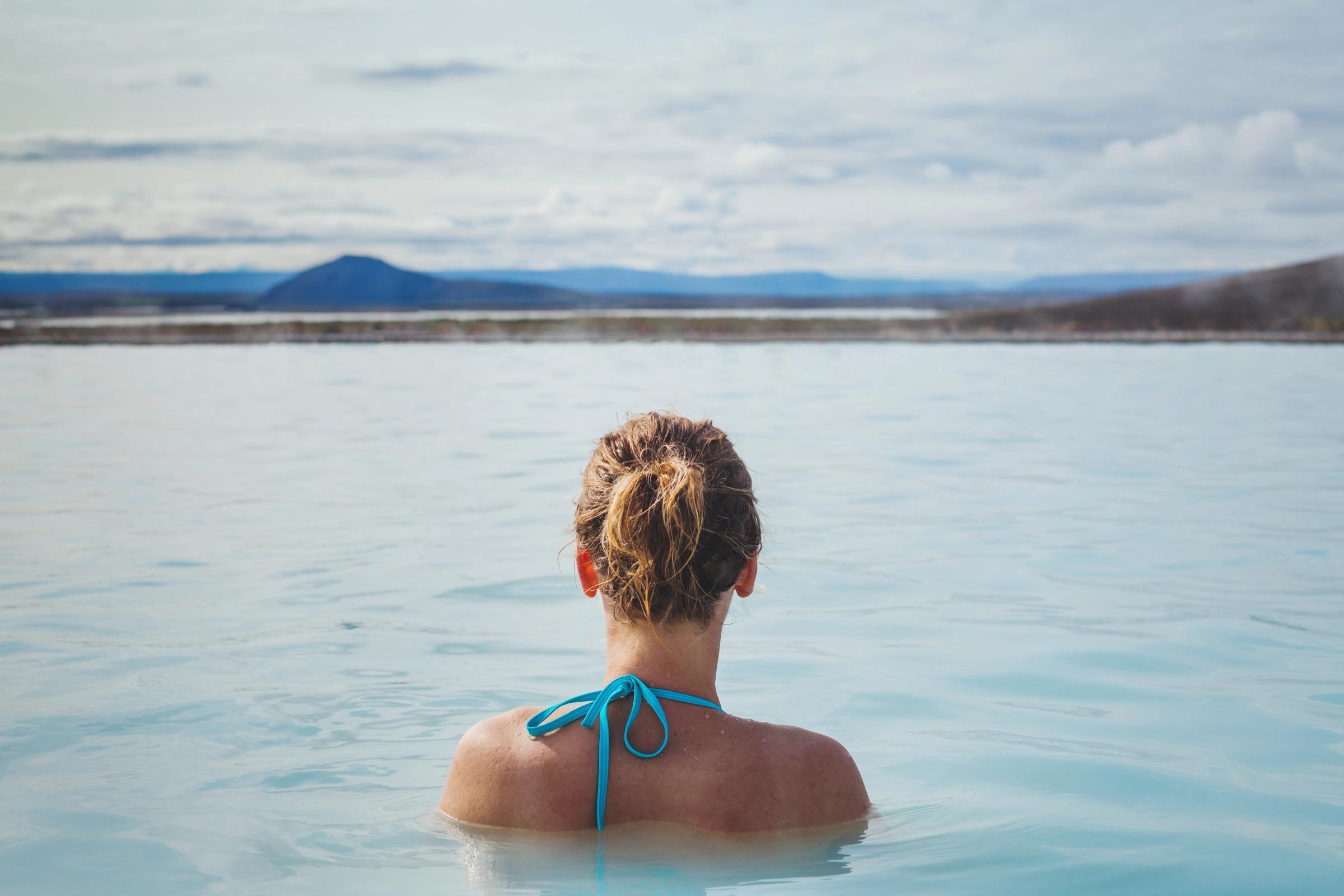 Espalda de una persona en una piscina geotérmica azul claro, mirando hacia unas montañas lejanas.