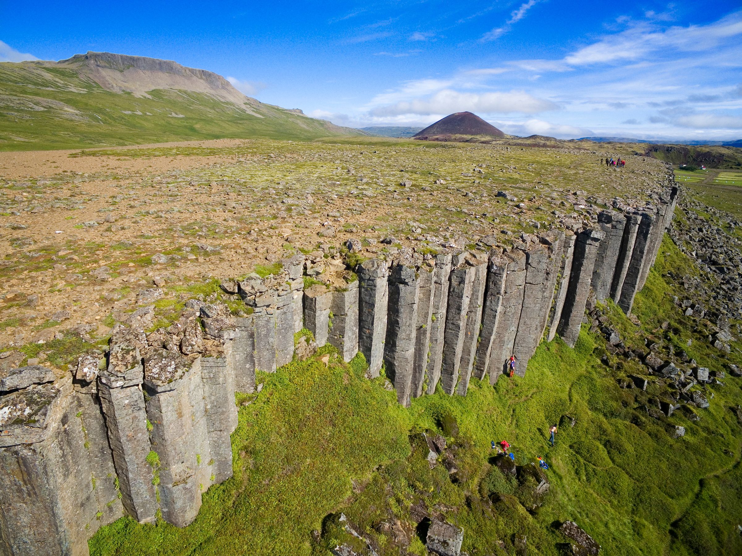 Basalt column cliffs with people hiking and climbing, a green landscape, distant mountains, and a conical hill.