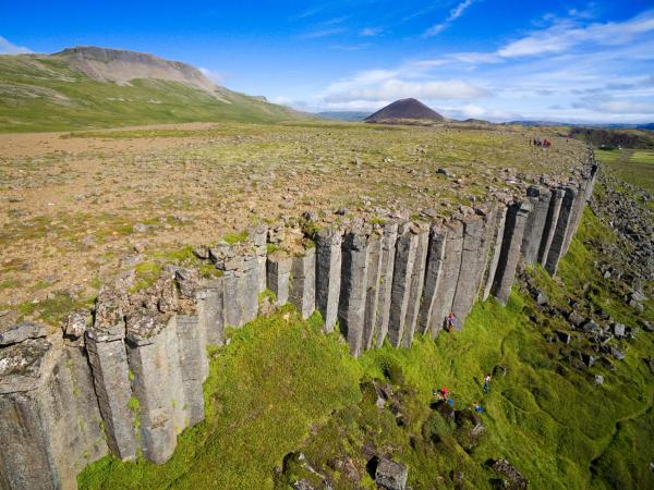 Basalt column cliffs with people hiking and climbing, a green landscape, distant mountains, and a conical hill.