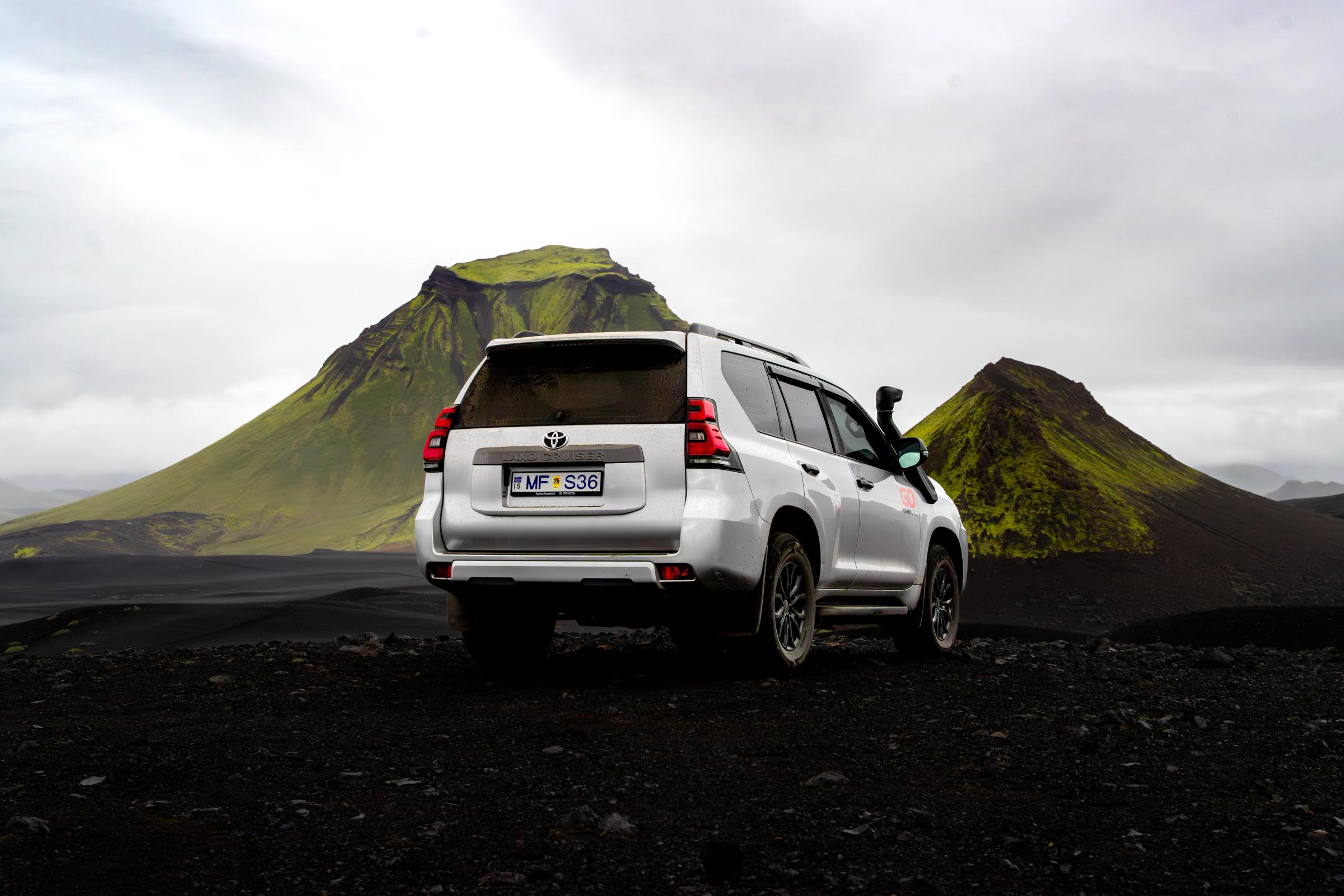 SUV plateado todoterreno sobre un suelo oscuro, con dos montañas verdes cubiertas de musgo al fondo.