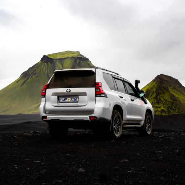 A silver SUV on black volcanic ground with two green moss-covered mountains in the background.