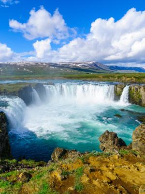 A wide waterfall cascades into a turquoise river, surrounded by green cliffs and distant mountains under a cloudy blue sky.