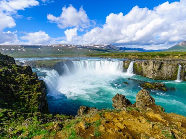 A majestic waterfall with vibrant turquoise water cascades over dark rocks, surrounded by green and rocky banks, with distant mountains and a bright blue sky with white clouds.