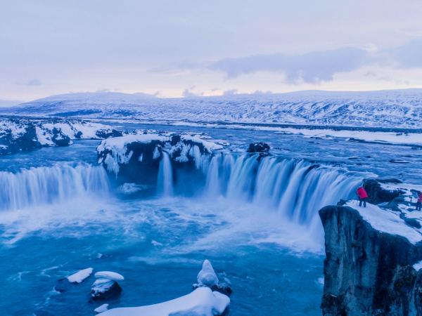 A wide waterfall in a snowy, blue landscape with people on a cliff.