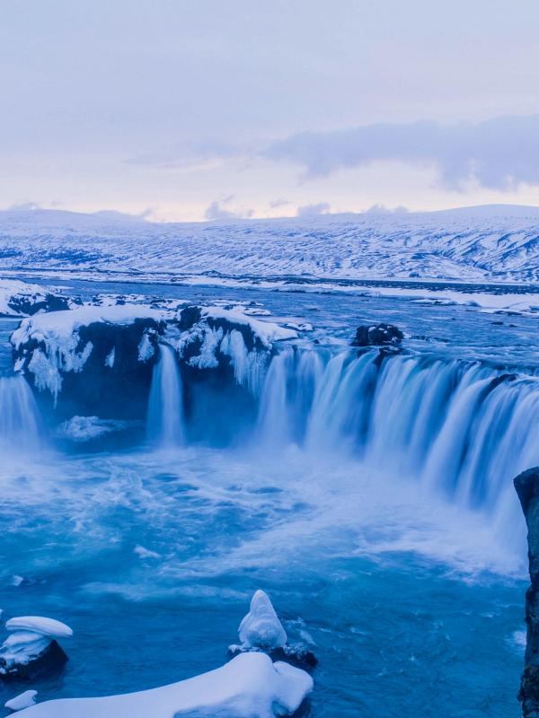 A wide blue waterfall in a snowy winter landscape with a few people on a cliff.
