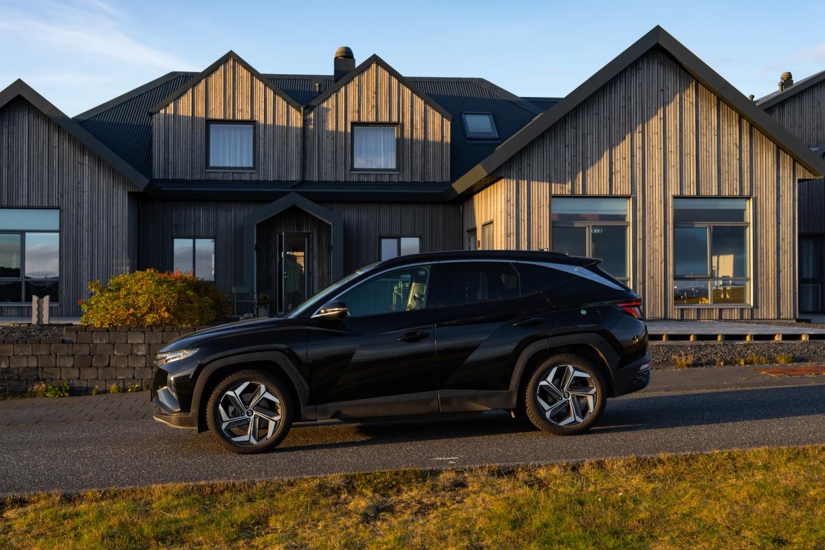 Vue de profil d’un Hyundai Tucson noir sur une route goudronnée devant une maison au bardage en bois