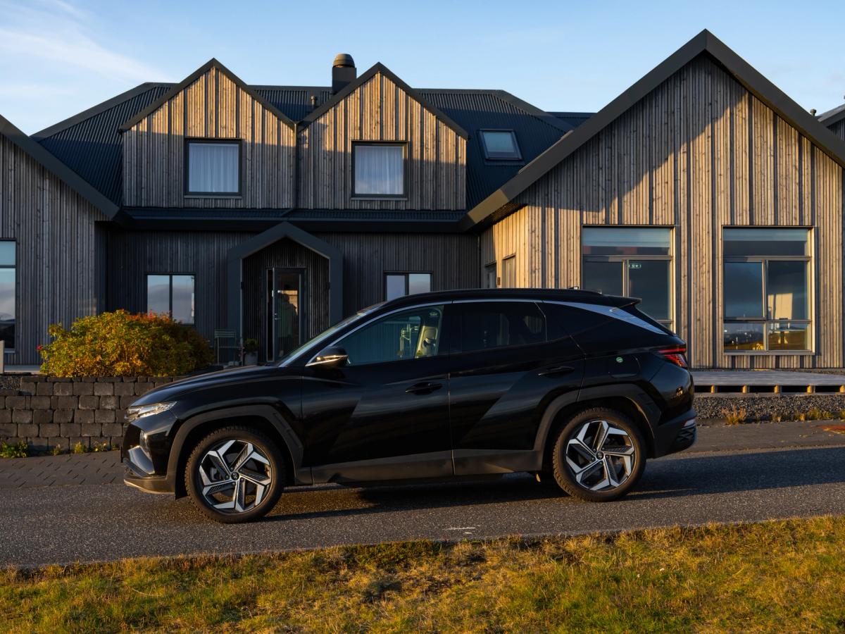 Vue de profil du Tucson Vue de profil d’un Hyundai Tucson noir sur une route goudronnée devant une maison au bardage en bois