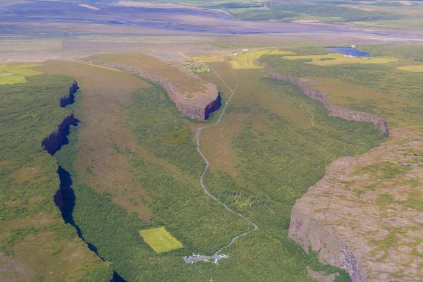 Aerial view of a deep, winding green canyon cutting through a vast landscape under a blue, cloudy sky.