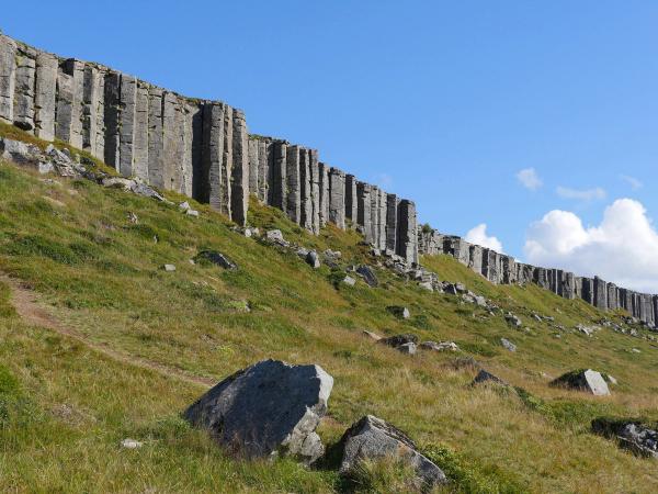 A long wall of grey basalt columns stretches across a green, rocky hillside under a blue sky.