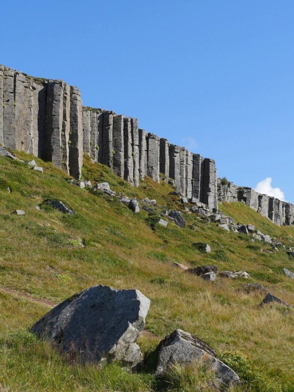 A long wall of grey basalt columns stretches across a green, rocky hillside under a blue sky.