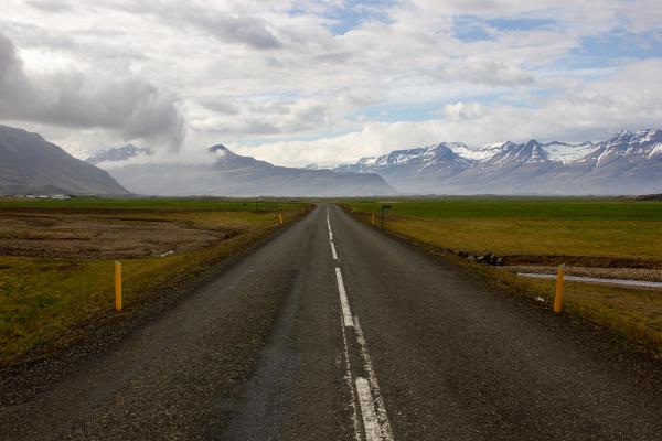 A straight road extends through green fields toward snow-capped mountains under a cloudy sky.