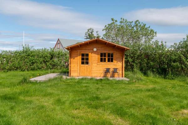 A small wooden cabin with two chairs in a green grassy field, with bushes and a house in the background.
