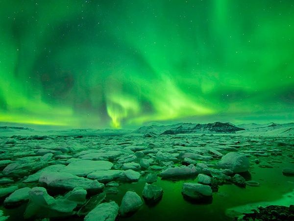 Vibrant green aurora borealis over an icy lagoon with distant mountains.