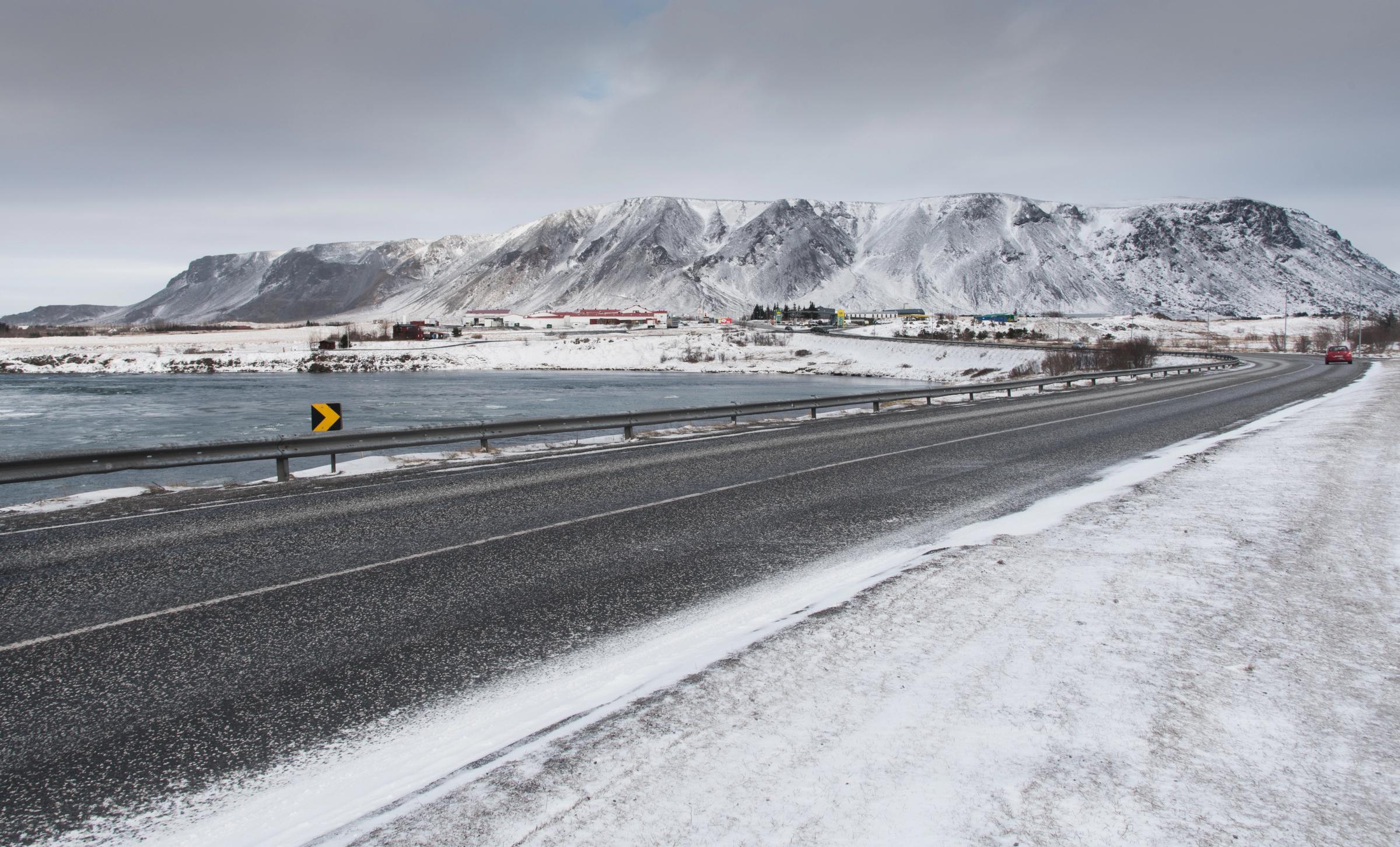 A curving road in a snowy landscape next to a lake, with mountains and buildings in the distance.