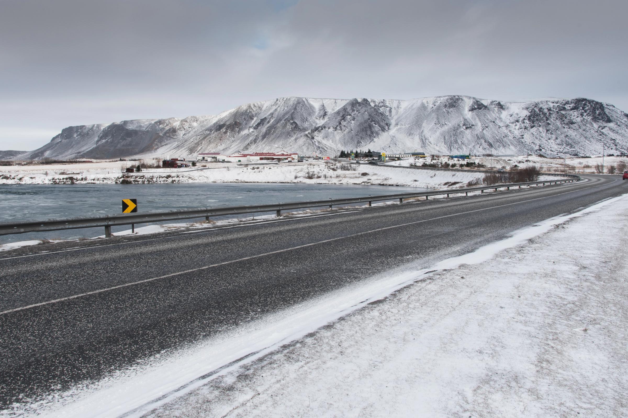 Paved road next to a body of water in a snowy mountain landscape with a distant town.