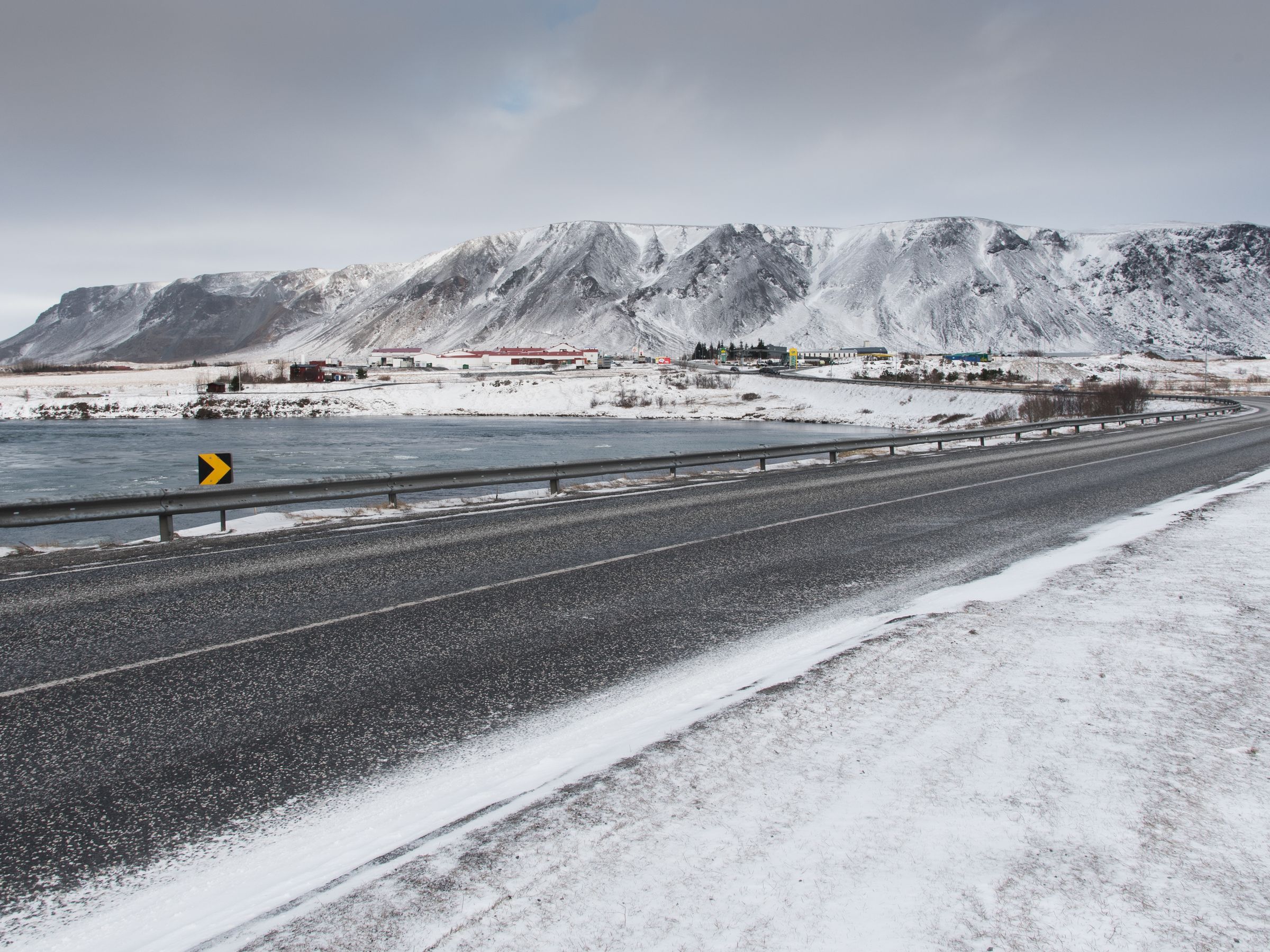 A curving road in a snowy landscape next to a lake, with mountains and buildings in the distance.