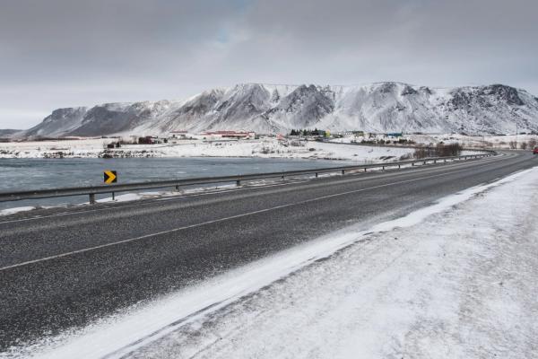 Paved road next to a body of water in a snowy mountain landscape with a distant town.