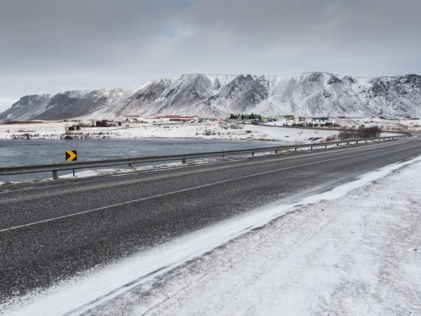 A curving road in a snowy landscape next to a lake, with mountains and buildings in the distance.