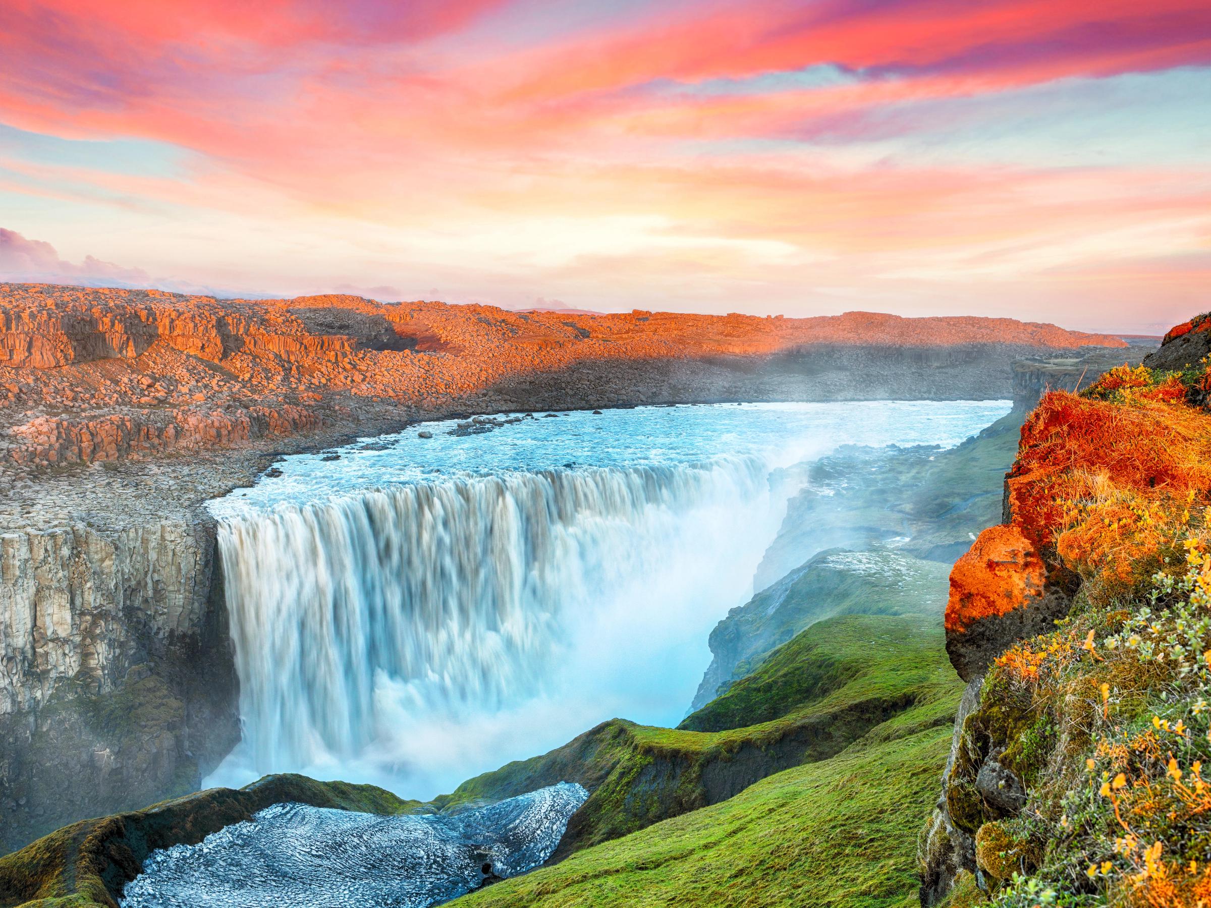 Una amplia cascada cae en un cañón rocoso bajo un cielo de atardecer vibrante, rodeado de acantilados verdes y cubiertos de hierba.
