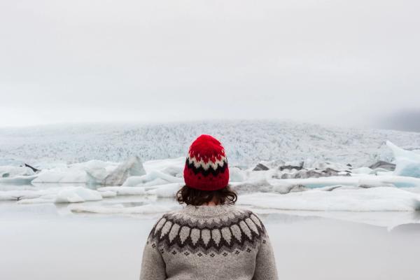 a woman in a red hat and sweater is standing in front of a glacier in Lopapeysa in iceland.