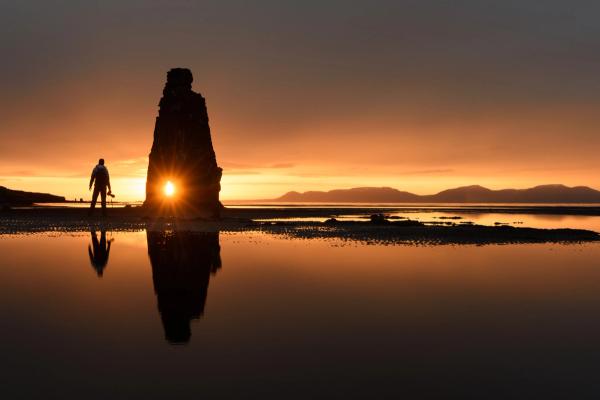 a man is standing in front of a large rock formation on a beach at sunset .