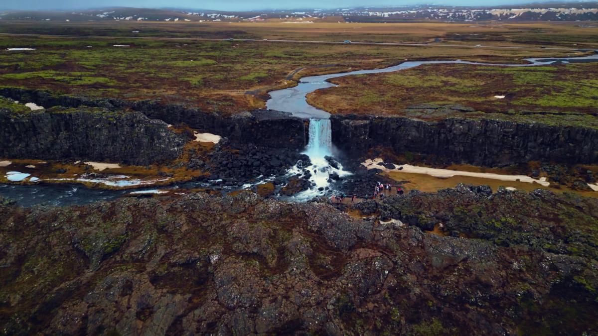 A waterfall plunges into a river through a dark rocky rift valley, flanked by green and brown landscape, with a winding river in the distance.