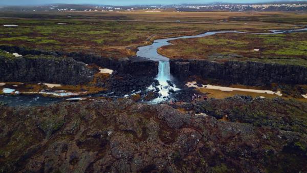 Aerial view of a waterfall cascading through dark rocky cliffs into a river, surrounded by a vast plain with green and brown vegetation.